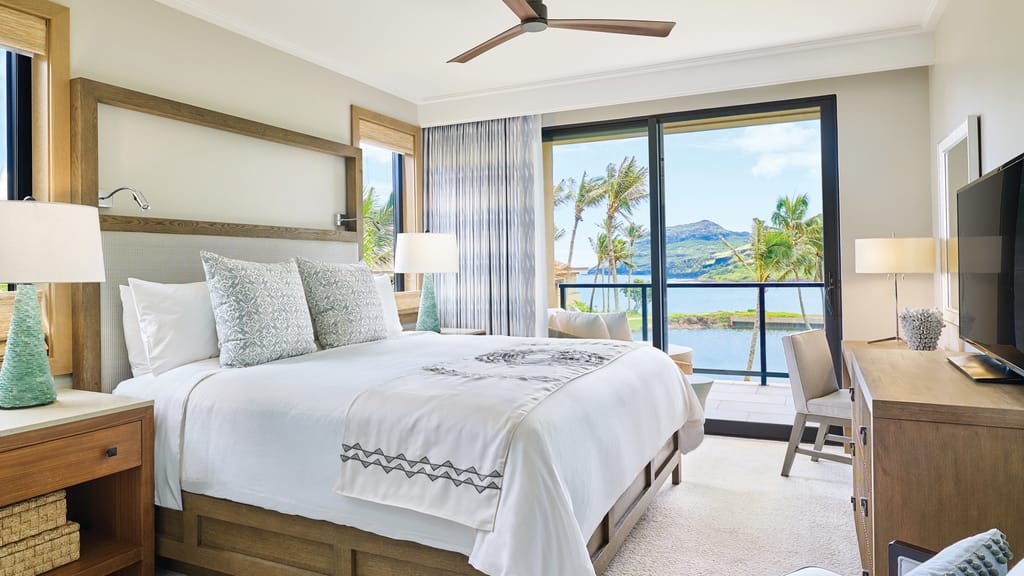 A wide-angle view of a hotel room with a large bed, desk, TV, and balcony overlooking the ocean.