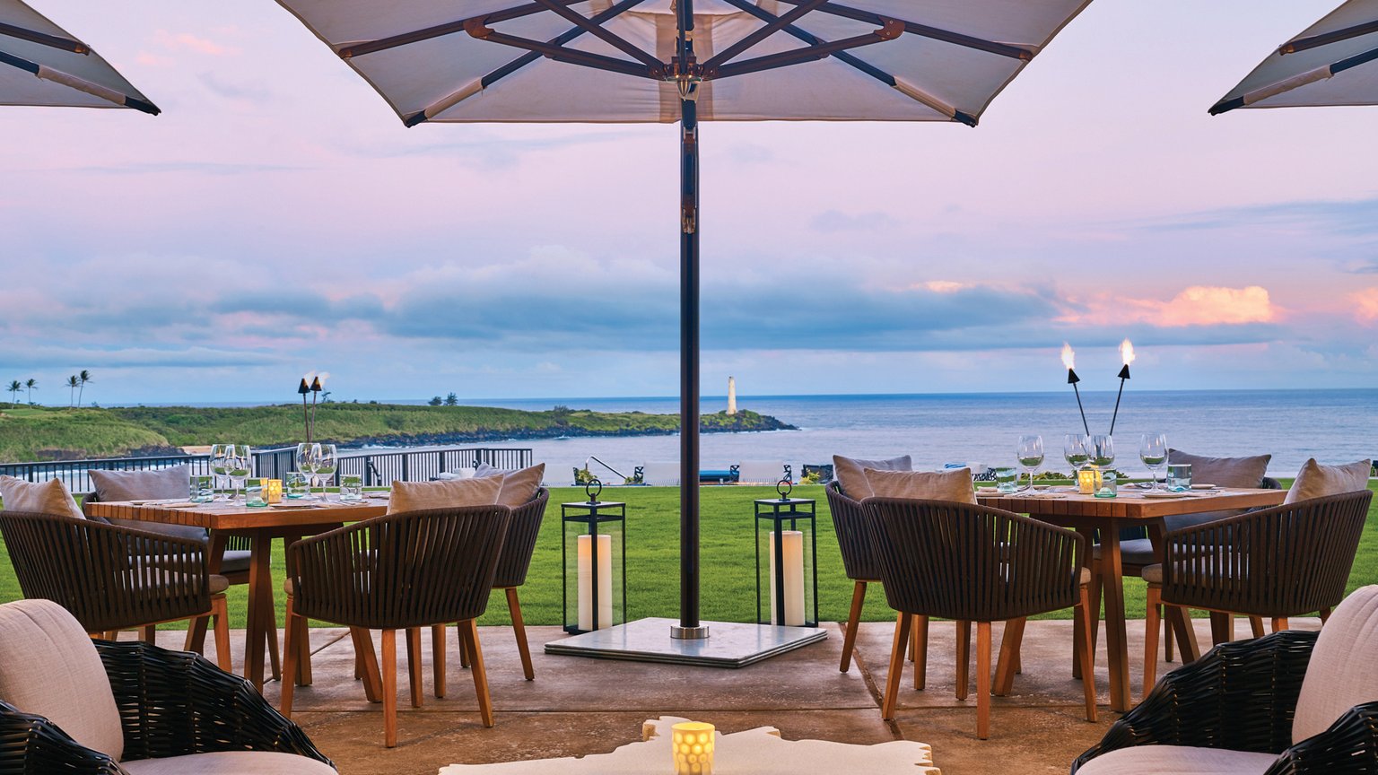 An outdoor dining area with a table set for dinner, overlooking the ocean at sunset.