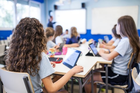 girl with brown curly hair studies in class with her tablet next to her classmates while they listen to the teacher